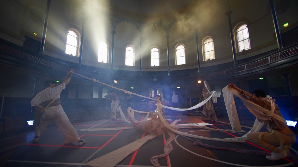 Photograph from a performance, in a dusty ound hall dancers pull sheets of fabric in a circle