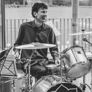 Robert is a white man with dark hair, in this photo sitting smiling behind a drum kit wearing a dark shirt.