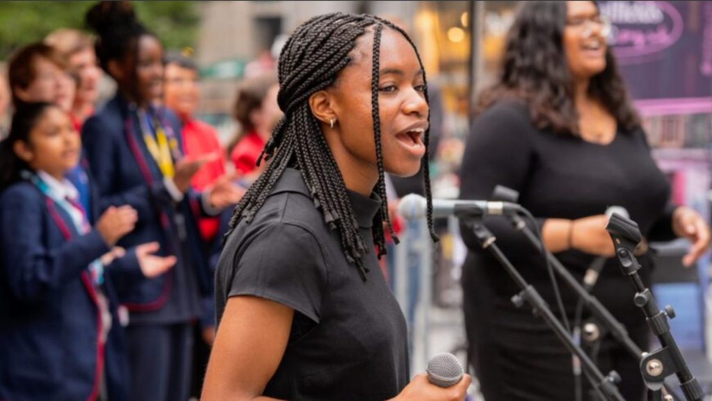 A young, black woman with braided hair is stood speaking in front of a microphone, onstage at some sort of outdoor, public event. There are a number of younger children in what appear to be school uniforms stood behind her onstage.