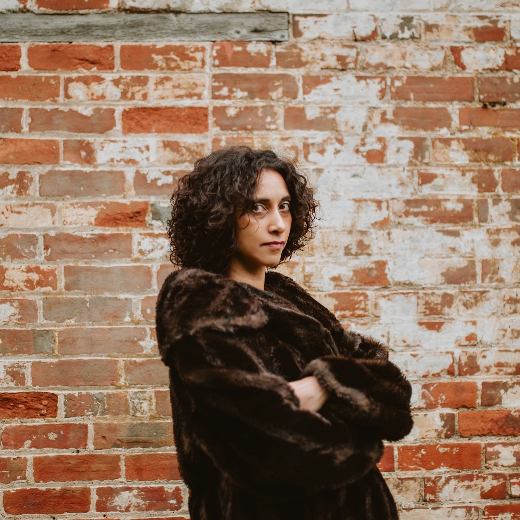Bethany stands sideways in front of a distressed red brick wall with her arms folded and a brown fur coat on. She looks sternly into the camera and her curly brown hair frames her face.