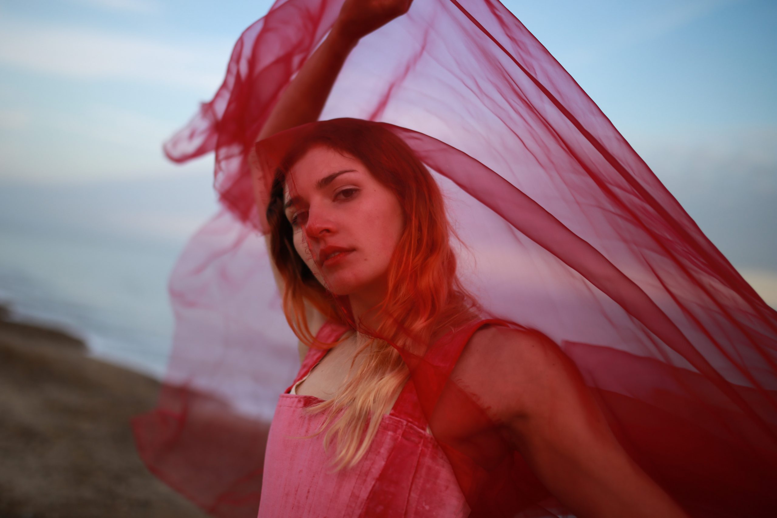 Samfire stands sideways to the camera which is angled up at her face. She drapes her face and upper body in sheer red fabric, holding it above her head. The out of focus background is coastal, with the sea and a small stretch of pebbles visible.