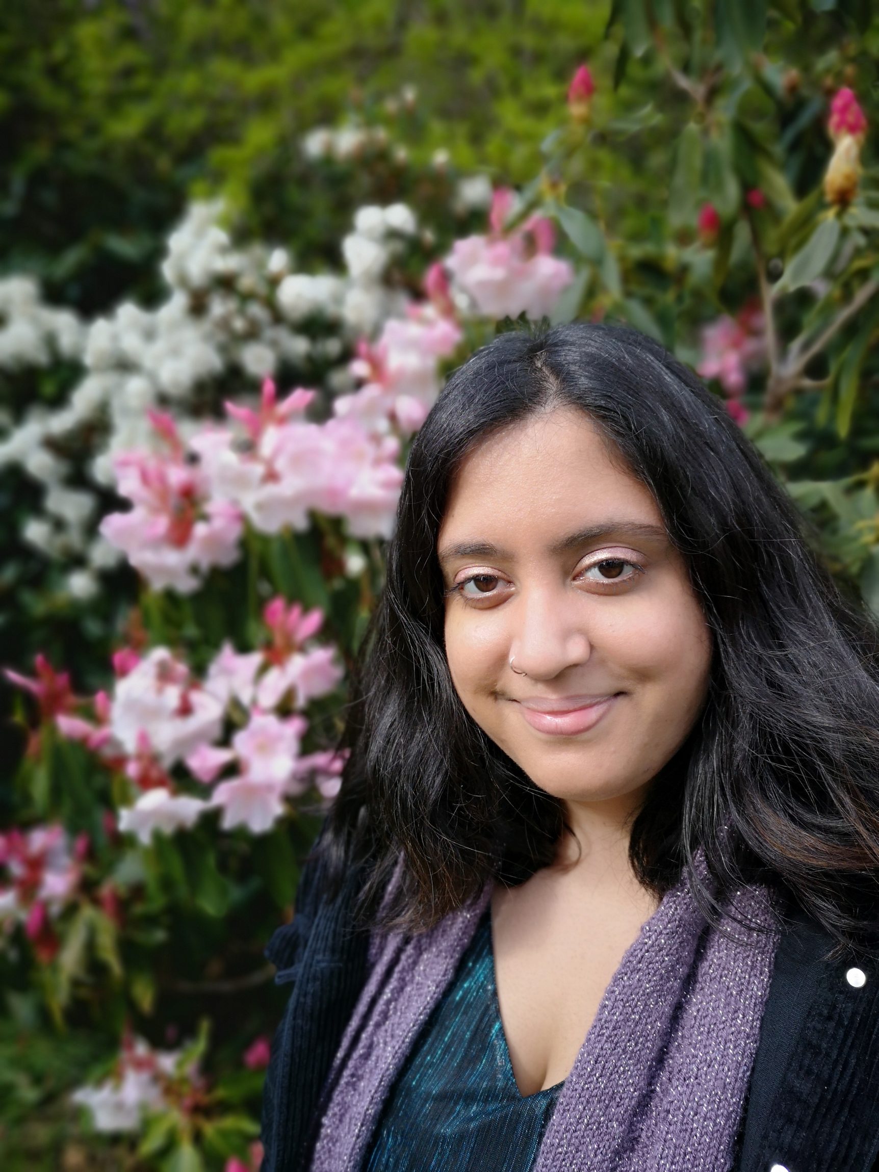 Composer Nikki Sheth faces the camera smiling in front of pink, red and white flowers and green foliage.