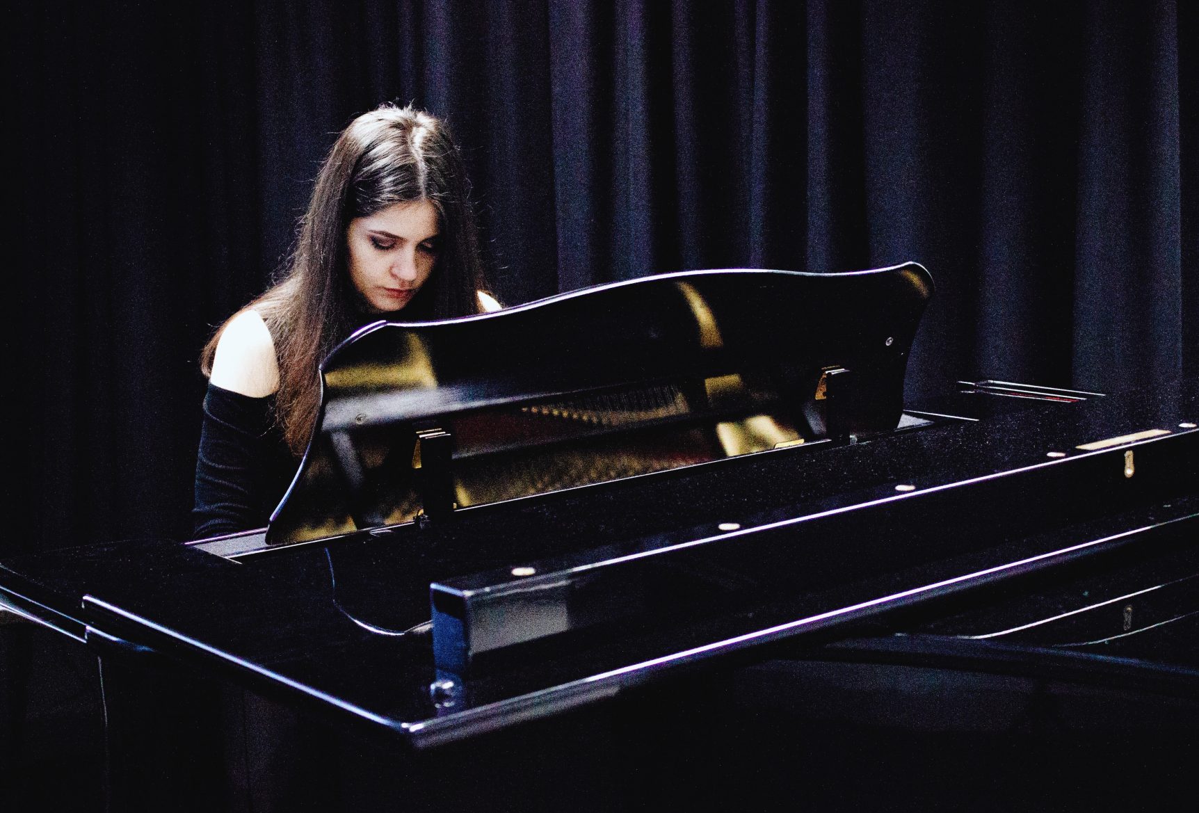 Adina sits behind a black piano in a room with black drapes. She looks down at the keys and her dark hair falls in front of her face.