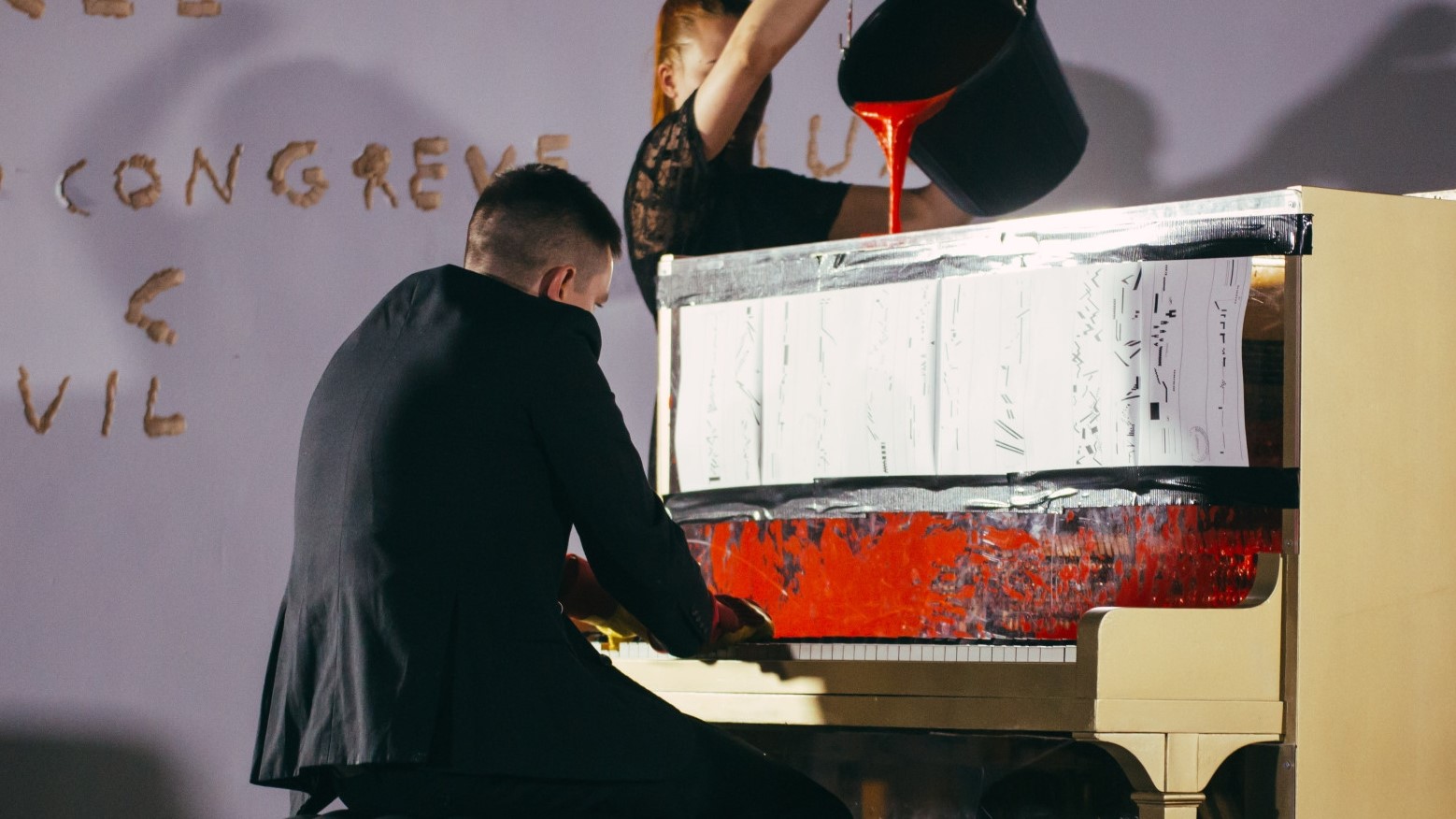 Andy Ingamells plays on a scruffy piano, while another person pours a bucket of red paint into its  workings.