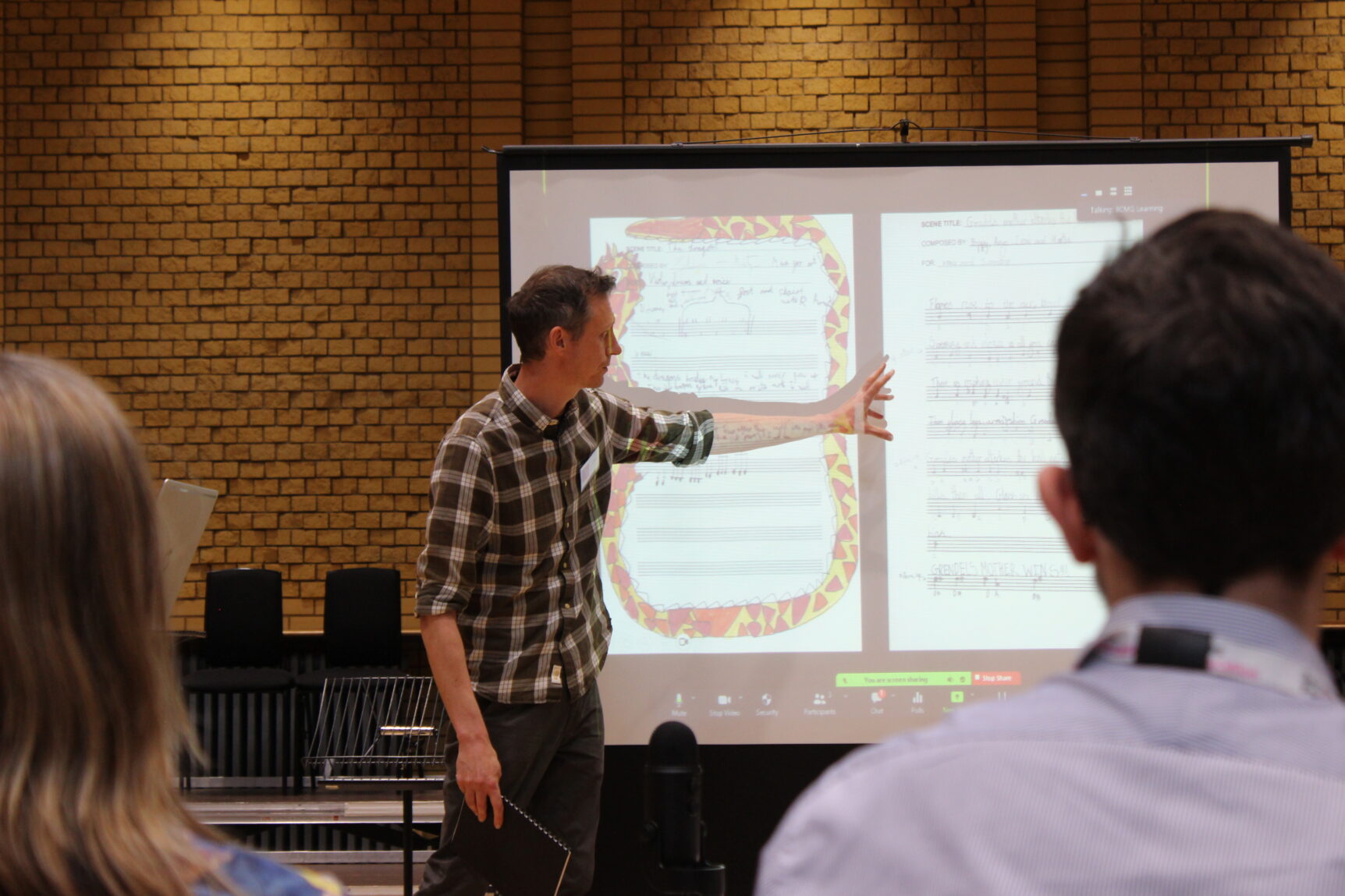 Composer Richard Bernard points to a white board showing an illustrated score. There are attendees in the foreground watching the presentation. The environment is the CBSO.