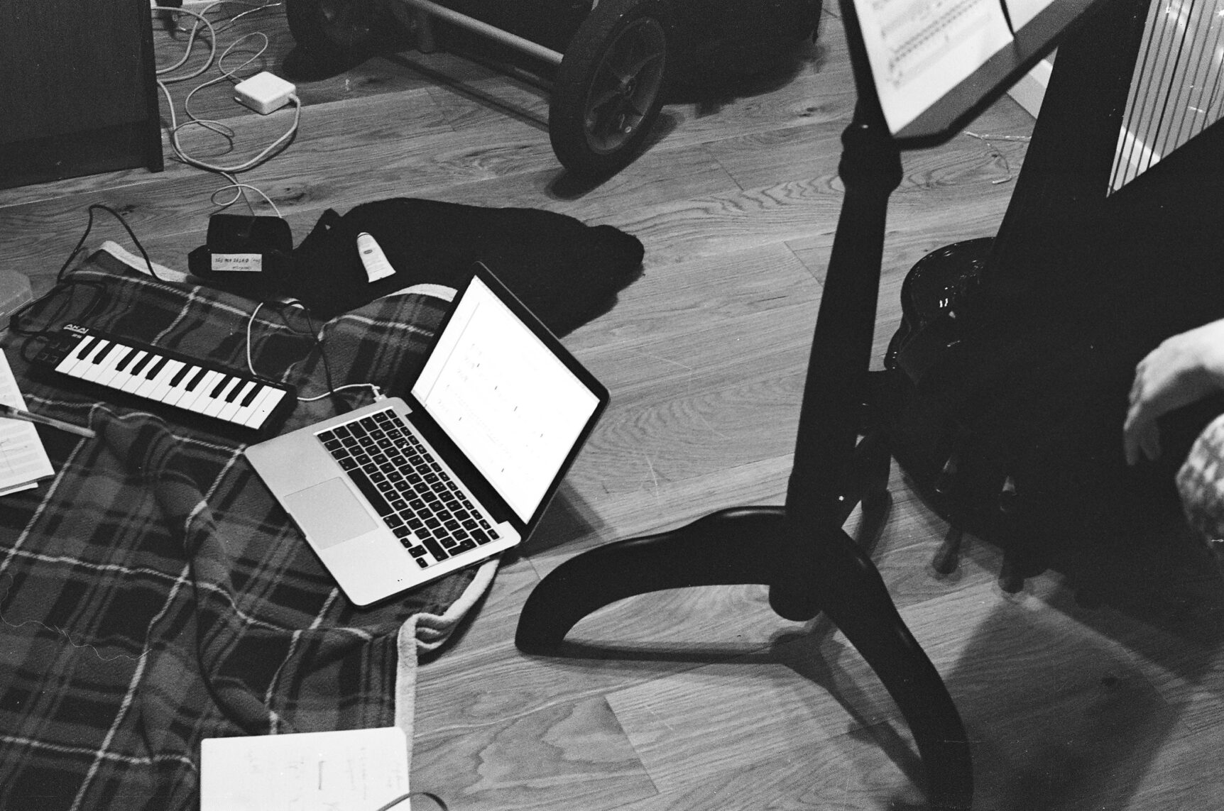 A black and white film photograph of a studio including a laptop and keyboard on a blanket on a wooden floor, and a music stand.
