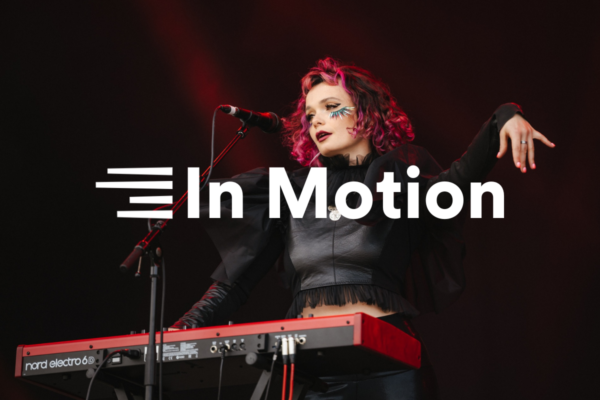 A photograph of composer Holysseus Fly performing with a microphone and keyboard. Text is overlayed reading “In Motion”