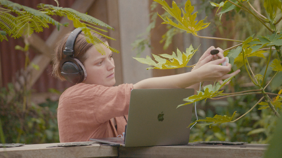A young woman holds up a recording device to a leaf while sitting at a laptop