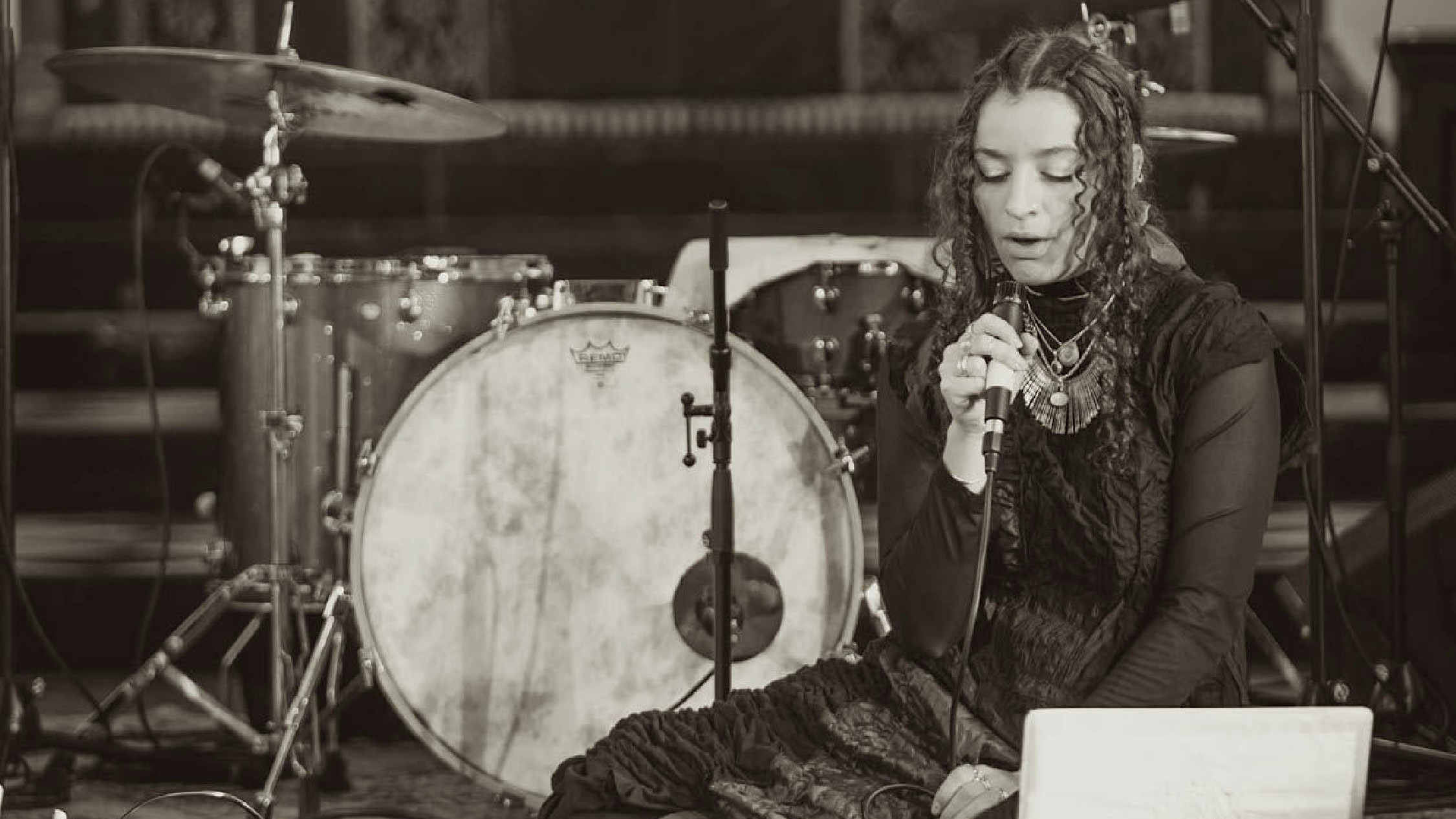 Composer Alliyah Enyo holding a microphone and performing, sitting in front of a drumkit